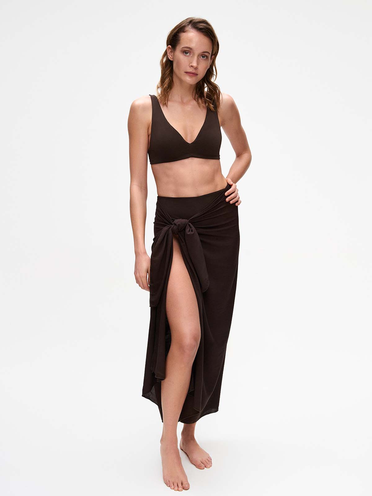 A woman wears the Kaiserin Selma Sarong – Coffee styled as a long wrap skirt with a front tie, paired with a matching brown bikini top, highlighting the lightweight viscose fabric and airy drape suitable for beach or resort wear.